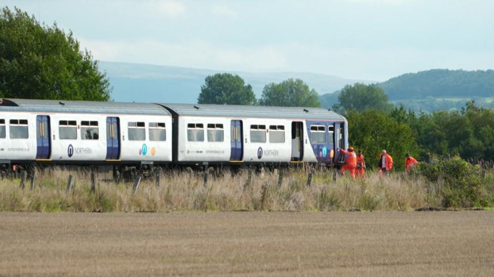 A Northern passenger train stands on a railway line in a rural landscape, with several rail workers in high-visibility orange clothing gathered beside the front carriage. Trees, rolling hills, and open fields surround the track under a cloudy sky.