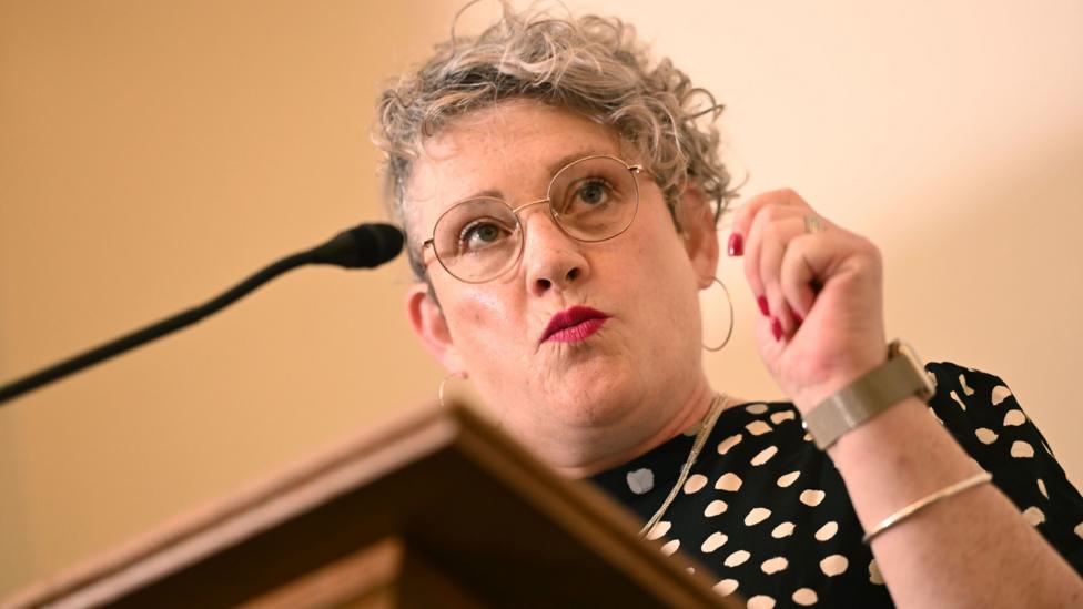 Ashley Dalton has curly blonde hair and wears a black dress with white polka dots. She is standing in front of a microphone at a lectern.