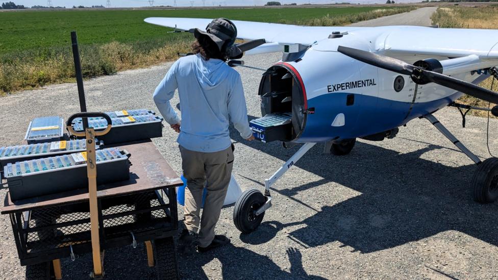 A Pyka drone on the runway has its nose removed while a technician swaps batteries.