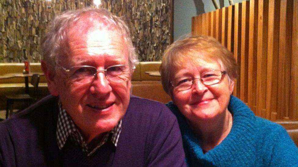 Two adults sit side by side in a restaurant booth, leaning slightly towards one another as they face the camera. The person on the left, Keith, wears rectangular glasses, a dark purple jumper and a checked shirt, while the person on the right, his wife Sue, wears glasses and a bright blue knitted sweater. The image is closely framed around the two seated figures.