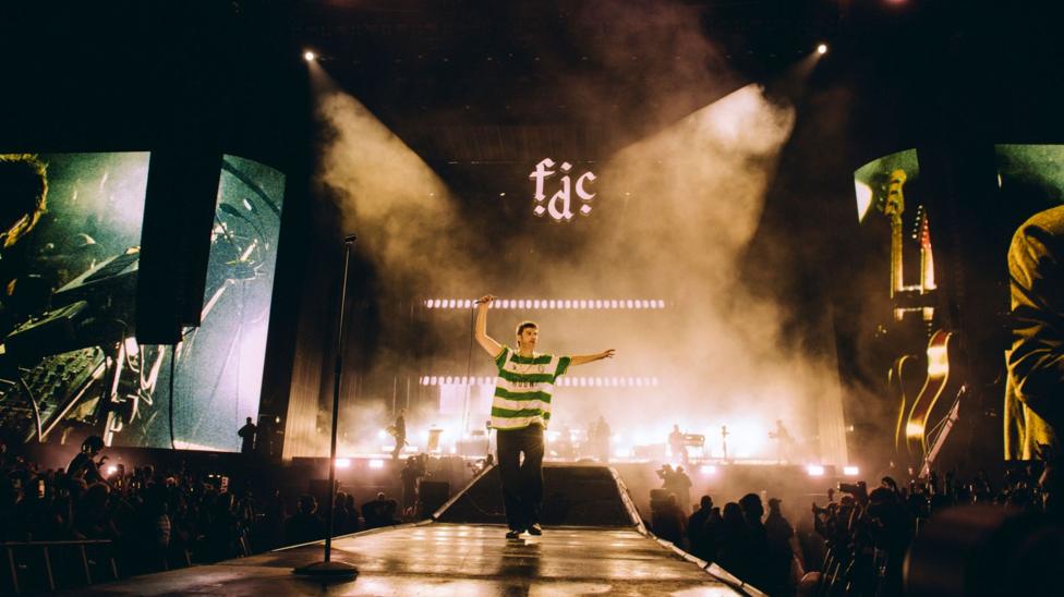 A landscape shot of Fontaines D.C. performing at Reading Festival. Grian Chatten is centre stage and seen on two large screens to each side of the stage, performing whilst wearing a Cwmbran Celtic FC away shirt.