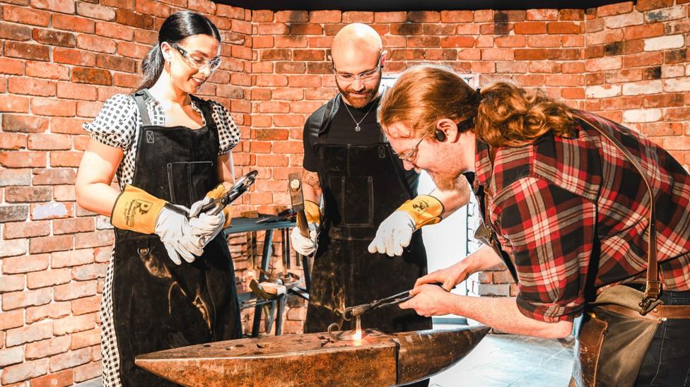 A man in a checked shirt with long red hair shows a couple how to work on metal at an anvil. The woman has long, dark hair pulled back while the man has a bald head and a red beard. They are both wearing gloves and black aprons.
