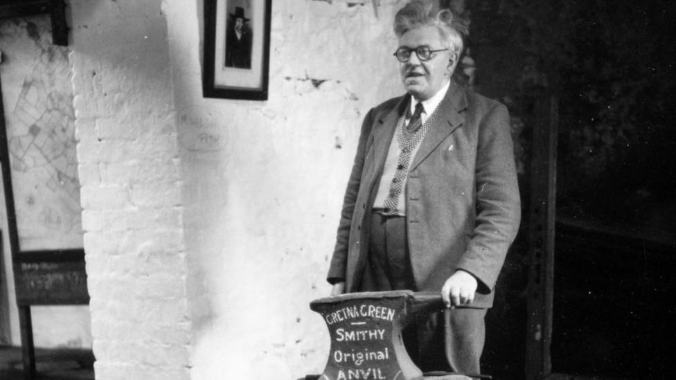 A grey-haired man in a jacket wearing glasses stands behind the Gretna Green anvil