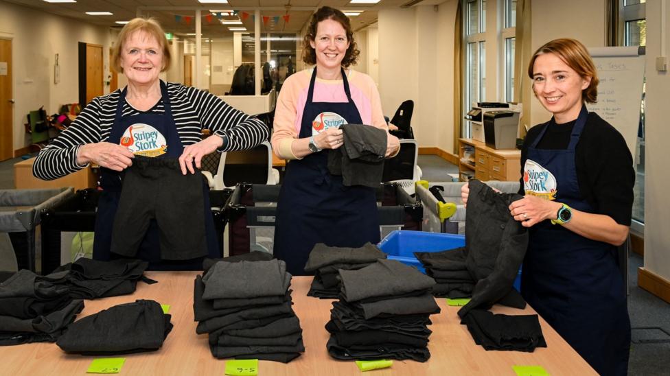 Three workers at Stripey Stork folding uniform items to be administered to children in Surrey. The three women are all wearing navy aprons with the charity's logo, and are all stood beside piles of grey uniform trousers. The lady on the left has blondish hair and is wearing a striped top. The lady in the middle has brown hair and is wearing a pink top. The lady on the right has light brown hair and is wearing a black jumper.
