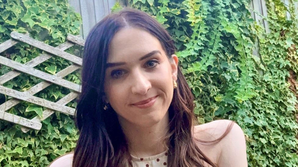 A photo of a woman with dark hair stood smiling in front of a hedge