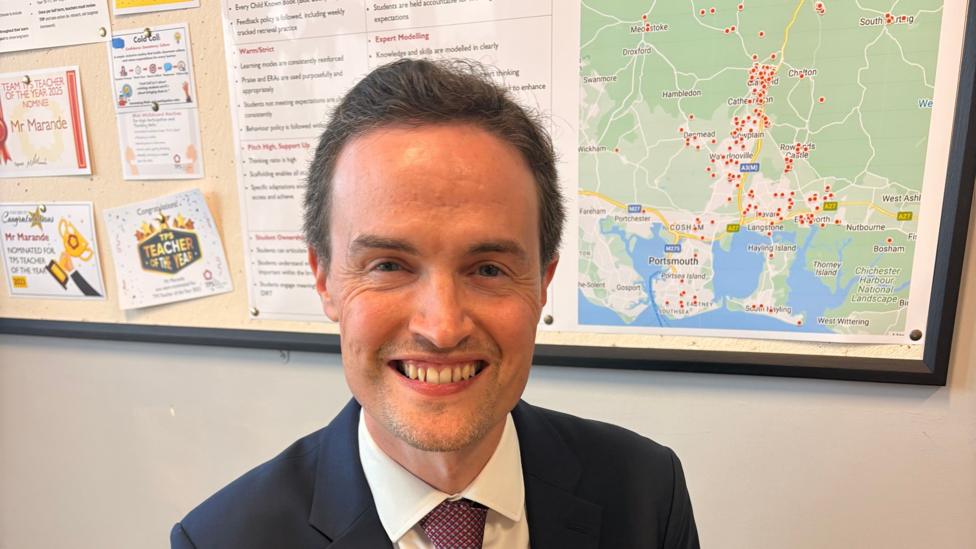 Mark Marande headteacher of The Petersfield School smiling, sitting in front of a pin board with a map and various notices on it.