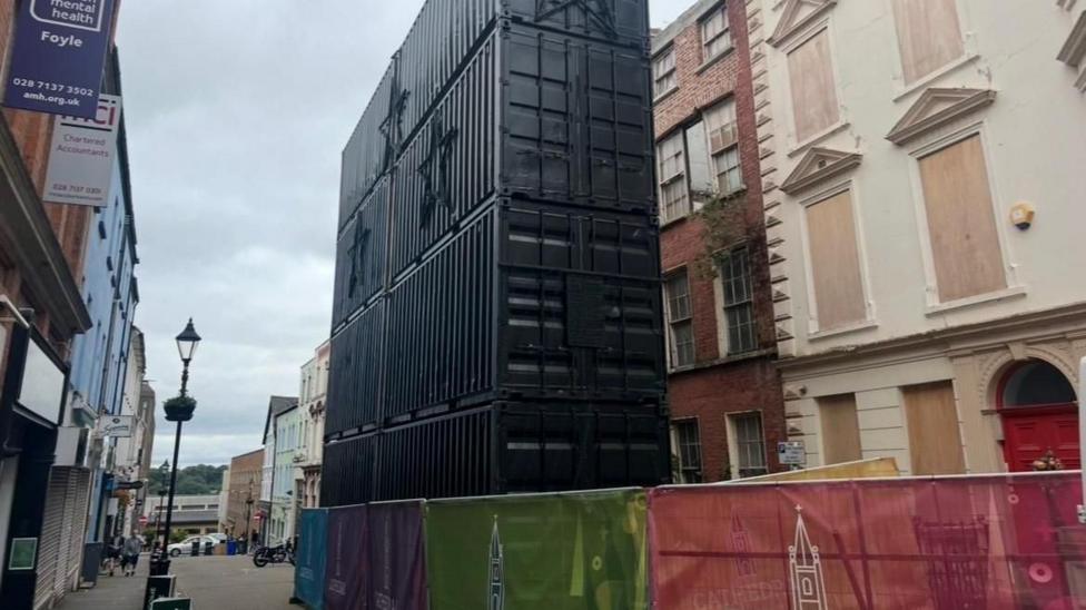 Large shipping containers stand upright in a narrow pedestrianised street. Shops line both sides of the street. A number of crash barriers are in front of the container.