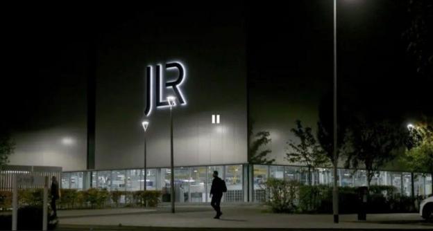 A little up sign, saying JLR, is on the side of a large, white warehouse-type building with ground floor windows all the way round its groundfloor level. The shot is taken at night and a silhouetted figure can be seen walking on the pavement outside.