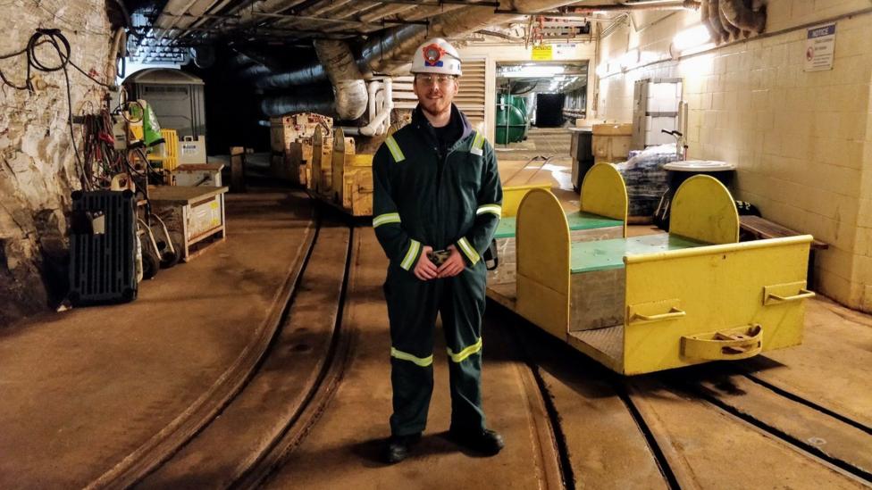 Dr Sam Eriksen smiles at the camera as he stands in an underground research facility. He wears a white hard hat with a head torch attached to it, and dark-green overalls with light green reflectors.
