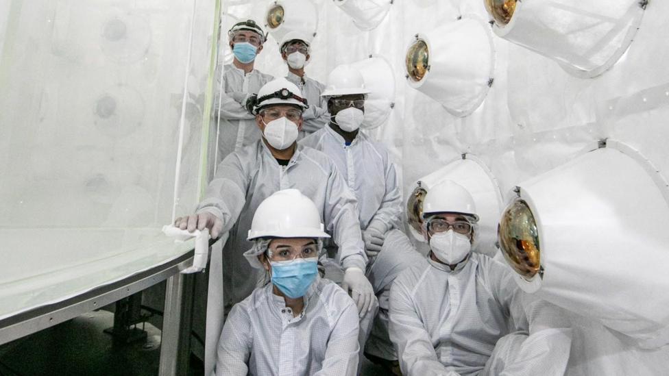 A group of scientists wearing white hard hats, masks, and white haz mat suits look at the camera as they are are gathered inside a water tank.