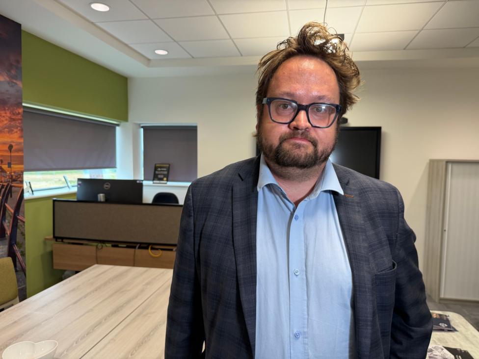Matt Storey is standing in a room, surrounded by light wooden tables and a desk with a computer in the background. He has dark messy hair and a beard. He wears glasses, a light blue shirt and grey jacket. He is looking at the camera with a serious expression.