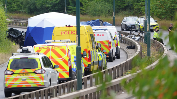 Police vans of varying size are lined up in a row leading up to the side of the wreckage, all with reflective liveries. To one side, two police officers rest with paper cups, with one sitting on the central reservation metal crash barrier that separates the two sides of the road.
