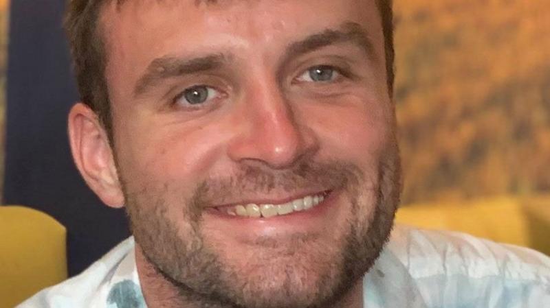 Alexander Harris, a man with brown hair and dressed in a light top, is smiling in a close-up shot