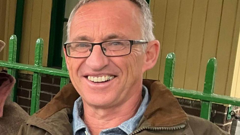 An older man with short white hair and glasses, wearing a brown coat, is smiling at the camera