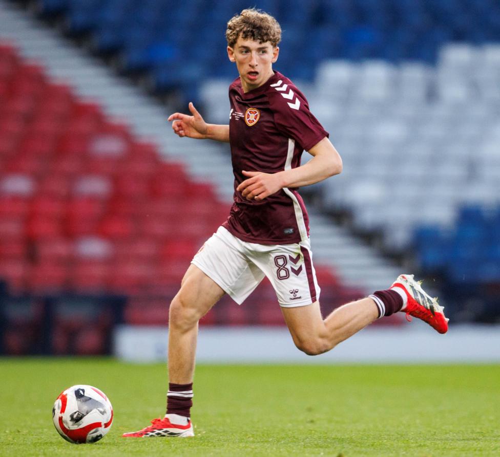 A young male footballer dribbling a ball at pace across a pitch. He has curly brown hair and is wearing a maroon Hearts strip.