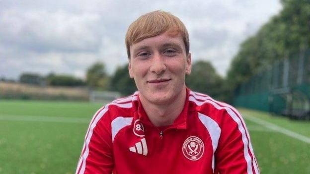 A young male footballer on a pitch looking into the camera and smiling - he has an auburn fringe and is wearing a red zipper with white stripes down the sleeves