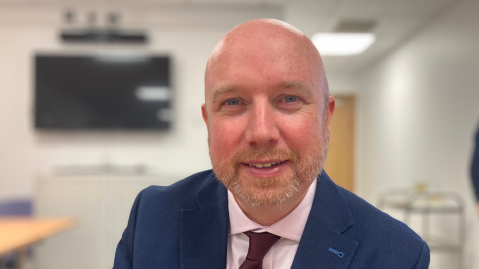 A bald man with a grey beard and moustache. He is sat down, wearing a blue suit jacket, pink shirt and red tie and is smiling as he looks directly at the camera.