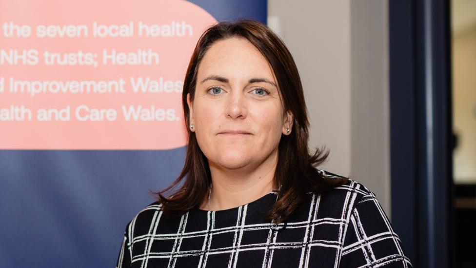 A woman with shoulder-length dark hair, wearing a black and white check top. She is stood in front of a red and blue Welsh NHS Confeseration branded banner and is smiling as she looks at the camera.