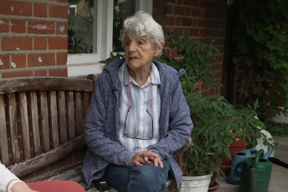 Katharine with white hair sits on a weathered wooden bench outside a red brick house, with potted plants and two green watering cans beside her.