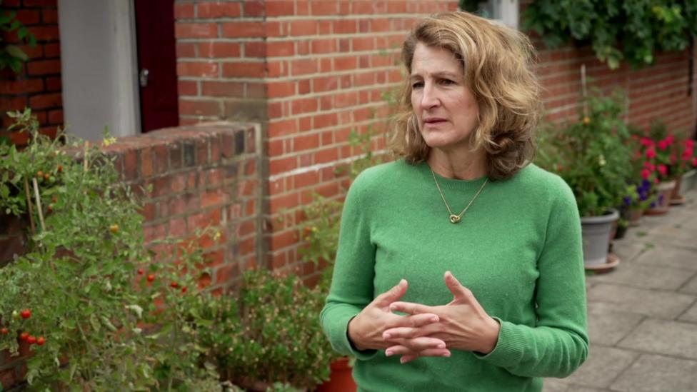 Neighbour in a green jumper stands on a pavement beside potted tomato plants and flowers, gesturing with both hands as she speaks