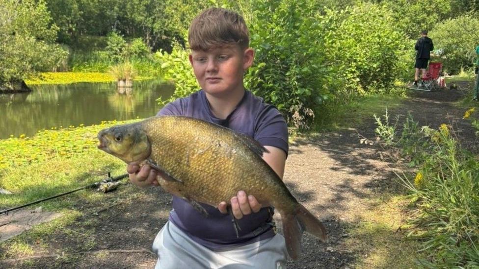 A teenage boy with light brown short hair is smiling while kneeling on grass by a river side while holding a large fish.