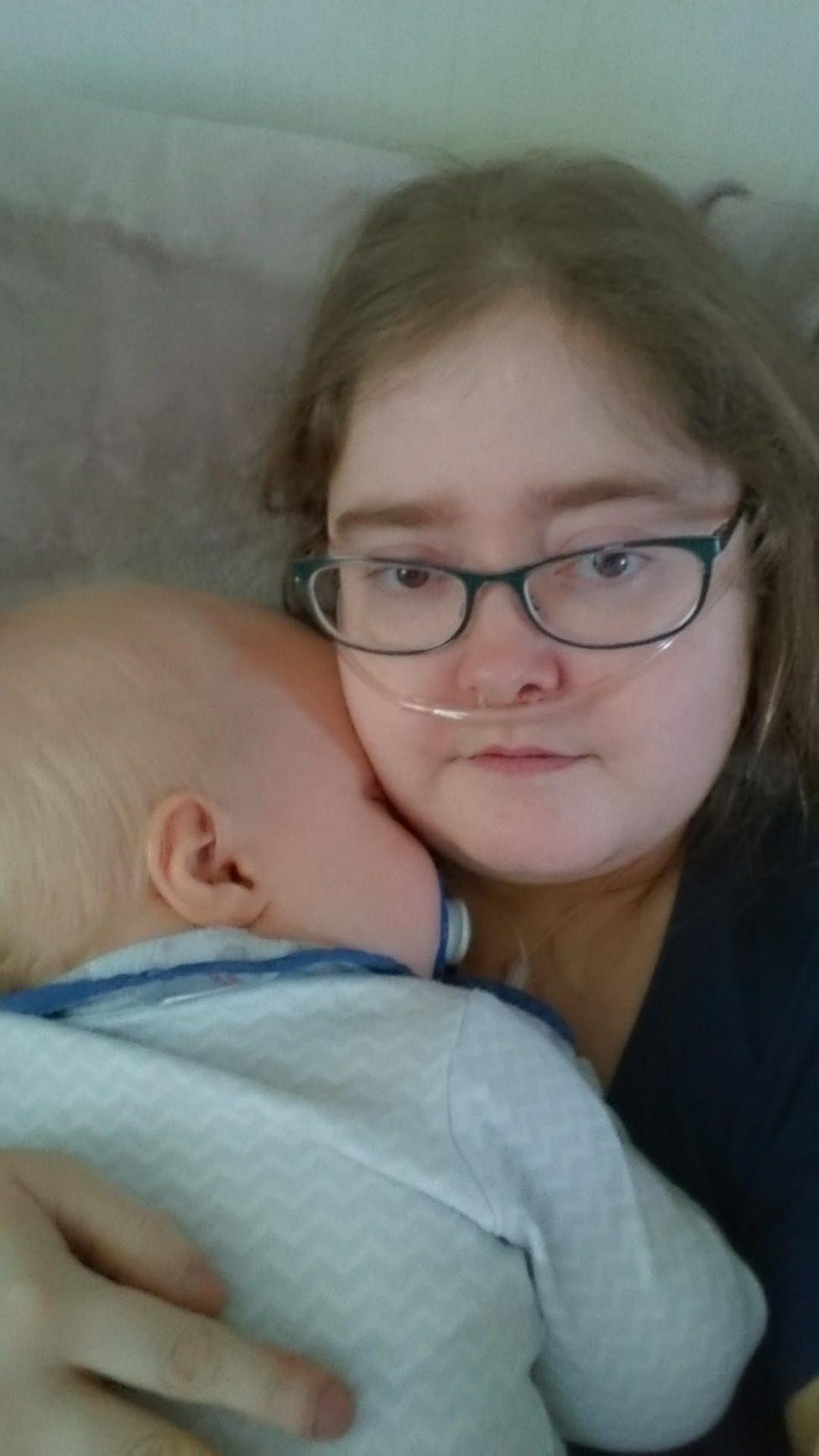 A woman with light brown hair and glasses, with a breathing tube, holds a baby