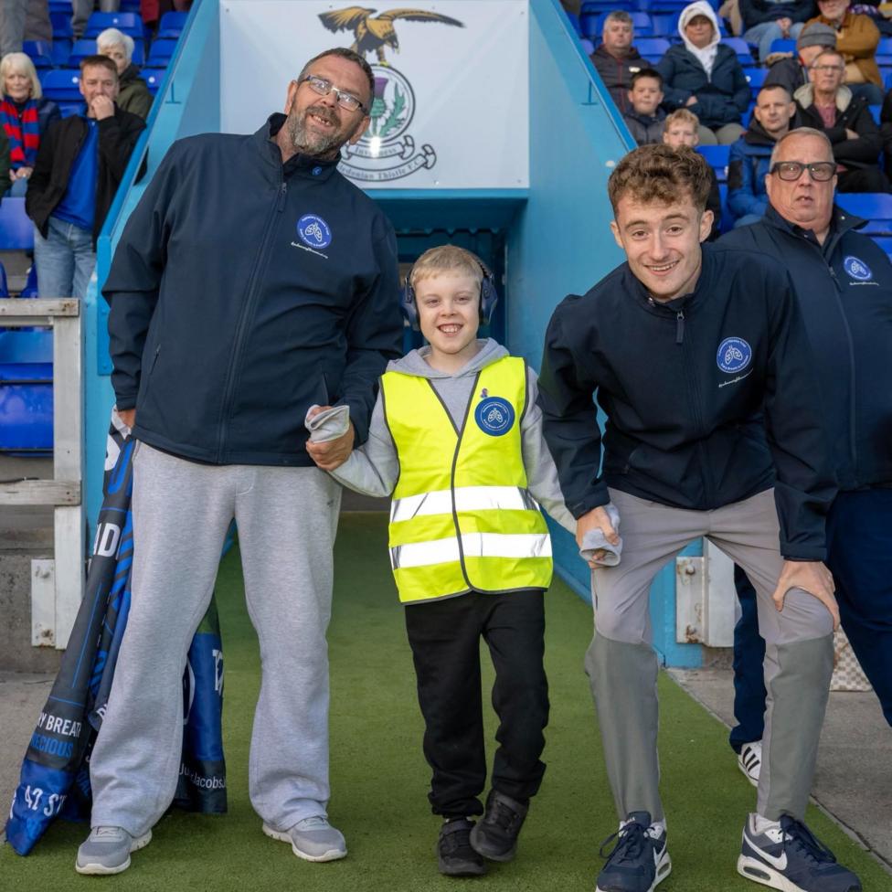 Three men and a young boy stand at the tunnel of a football ground with supporters in the background
