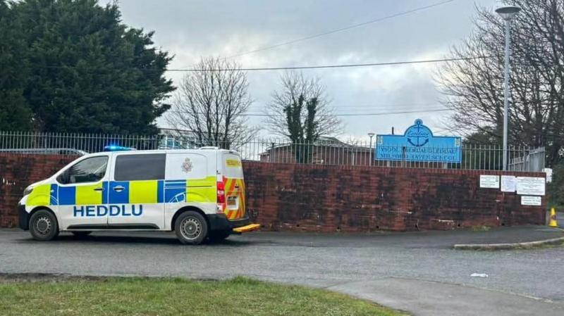 A brown school wall with a silver railing above and a police van parked outside. In the background is a blue school sign.