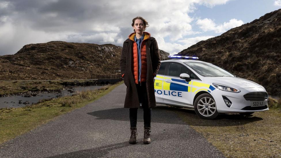 Sorcha Groundsell, a star of the show, stands in front of a police car on a single-track road. She is wearing a long coat over a red jacket and blue shirt and is wearing skinny jeans and boots. There are low, rugged hills behind her.