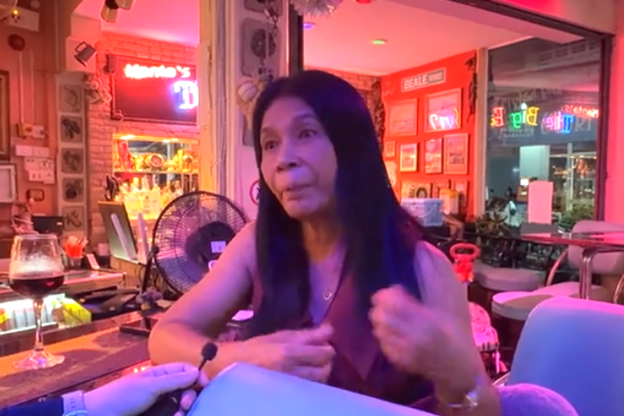 A middle aged Thai woman with long black hair, wearing a red vest, leans of a bar counter as she speaks into a microphone. She's inside a lowly-lit bar