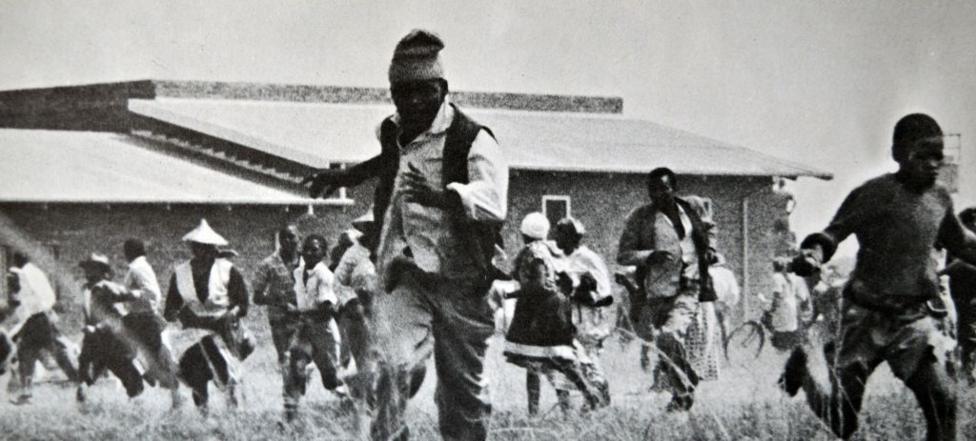 A grainy black & white image of a group of people running towards the camera. A low-rise building can be seen behind them.