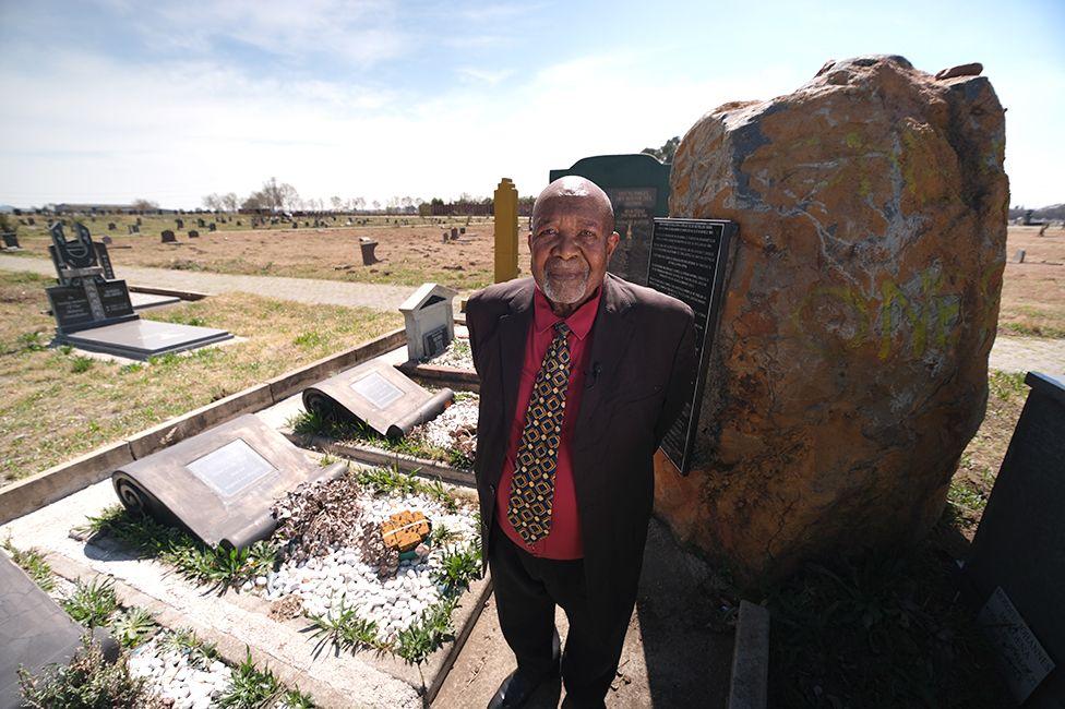 Larazus Magotsi in a dark suit, red shirt and tie stands by a large rock and graves.