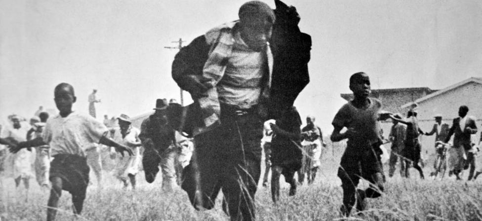 A grainy black & white image of a group of people running towards the camera. In the front is a man holding up his jacket - just behind him, either side, are tow children.