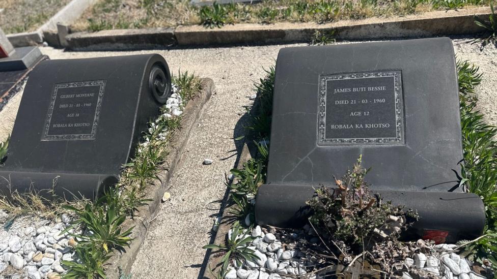 Two gravestones with weeds growing nearby among large pieces of gravel.