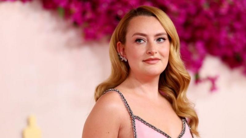 Amelia wears a bejewelled pink dress with straps. She stands against a pink floral background and looks to her side, with her strawberry blonde hair sitting at shoulder length