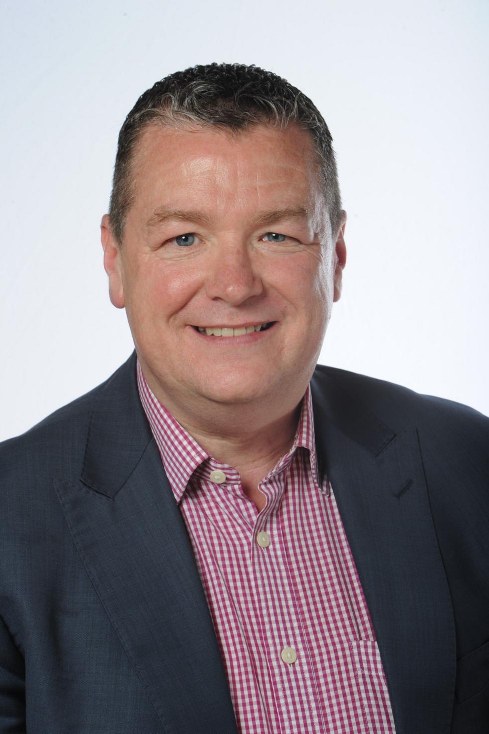 Ricky Bell poses for a formal head-and-shoulders portrait against a plain light background, wearing a dark jacket over a pink checked shirt and looking directly at the camera.