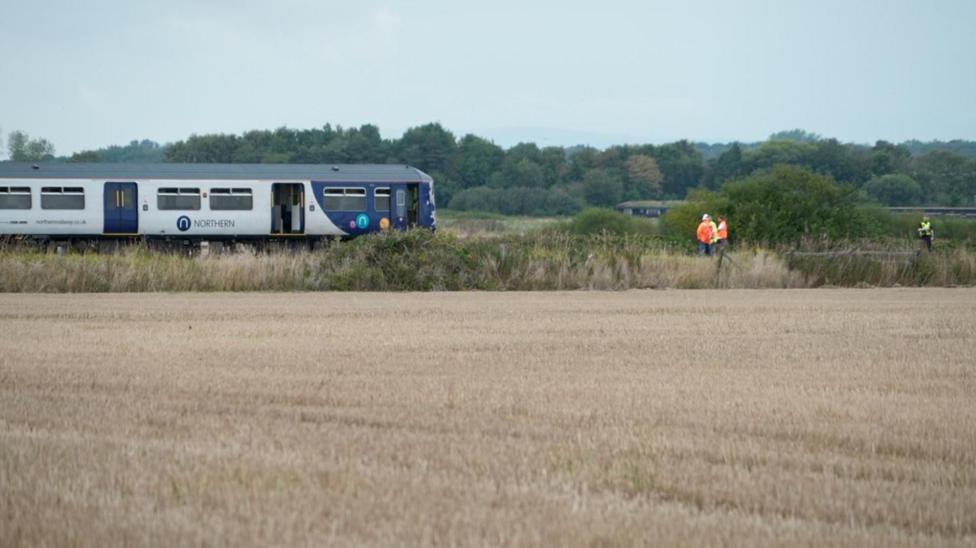 Image shows the Northern train halted on the railway line, with men in high visibility jackets walking on the track in front of it. The line is surrounded by fields.