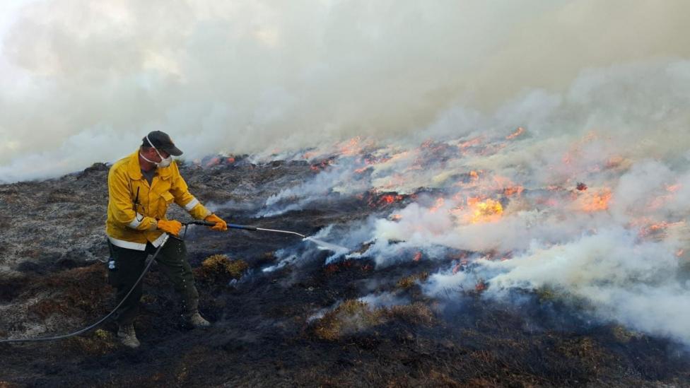 A man wearing protective clothing, a face mask and cap uses a small hose to put out flames in an area of peat.