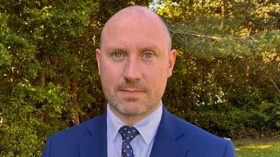 Neil Grey smiles at the camera. He stands outside in a garden and wears a dark blue suit with a light blue shirt and dotted navy tie.