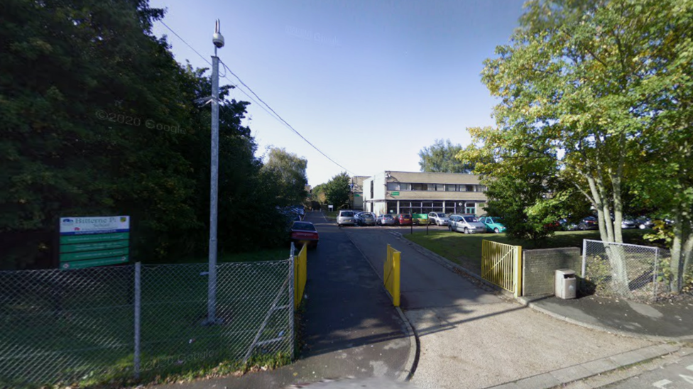 A two storey school building is pictured in the background, and in front of it are cars parked. The gates of the school are also pictured.