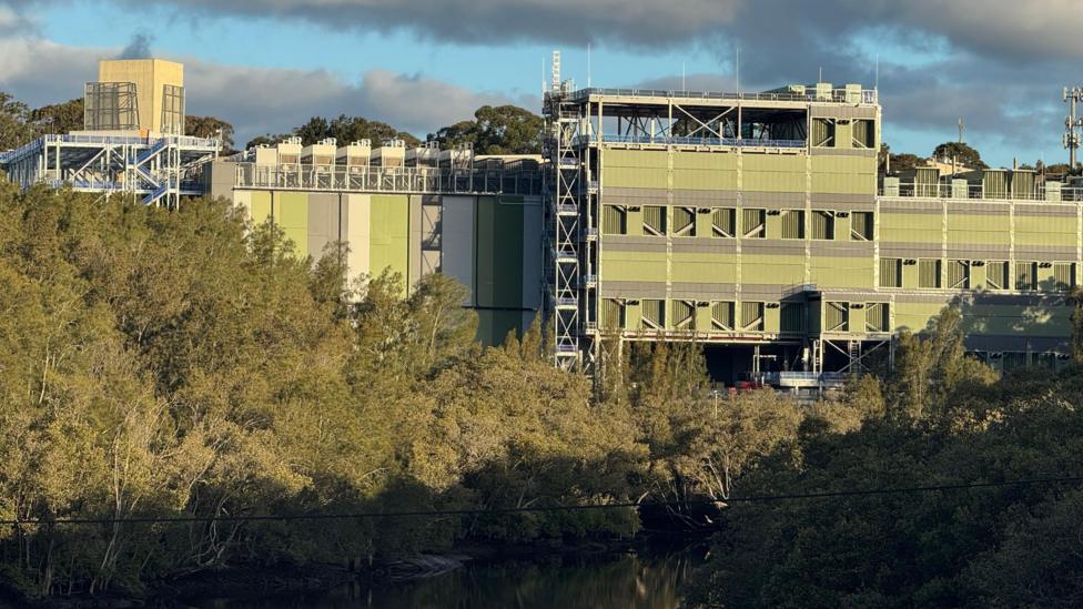 A large green and grey building situated amid bushland and a river down below.