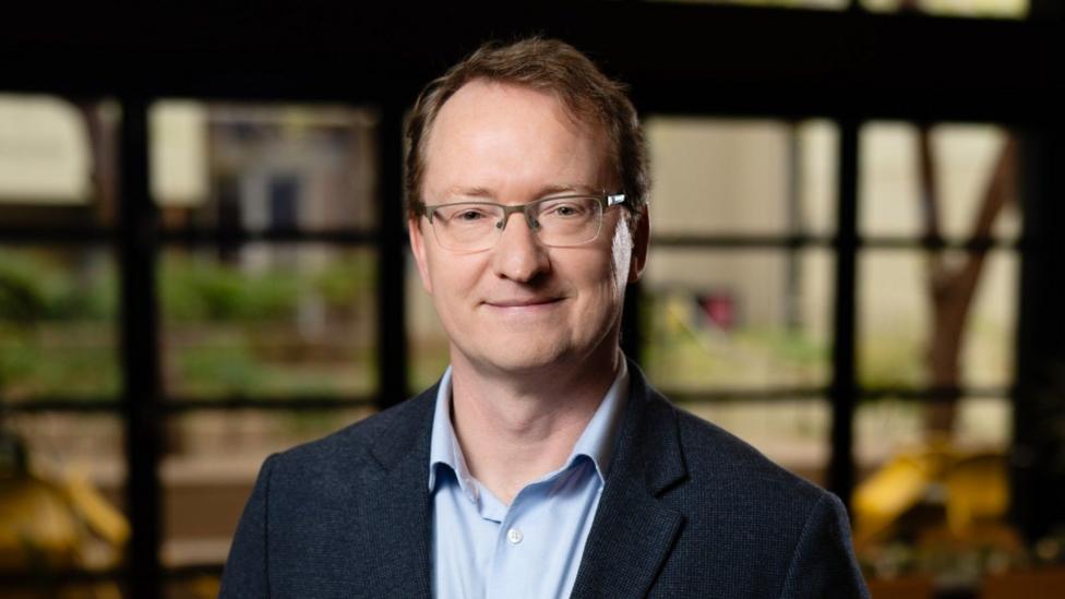 A middle aged man with short brown hair and glasses wearing a blue shirt and jacket smiles at the camera