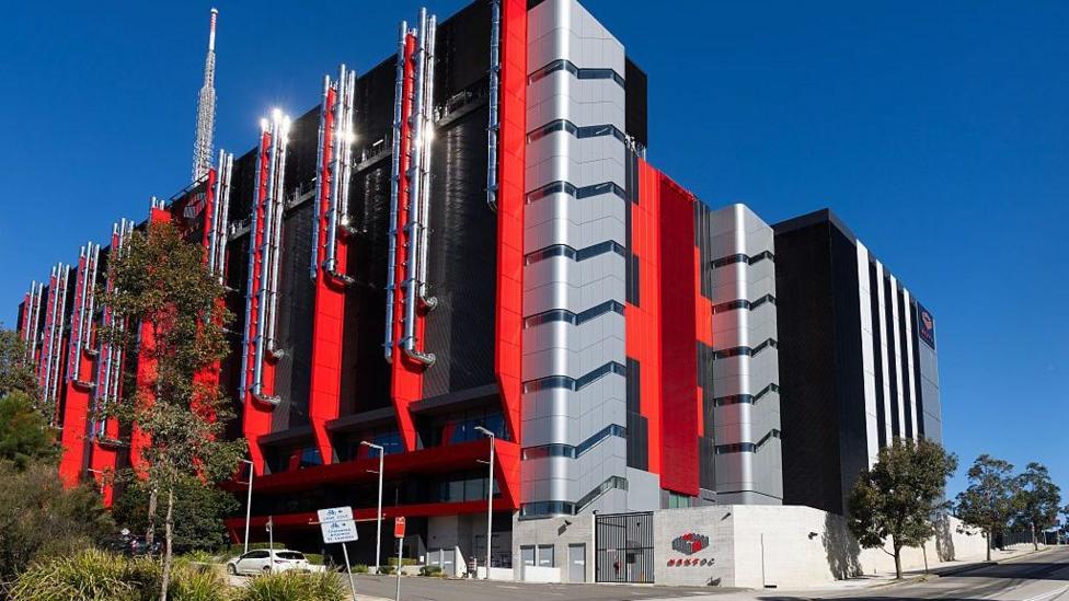 A huge red and grey windowless building with pipes on the outside against a blue sky.