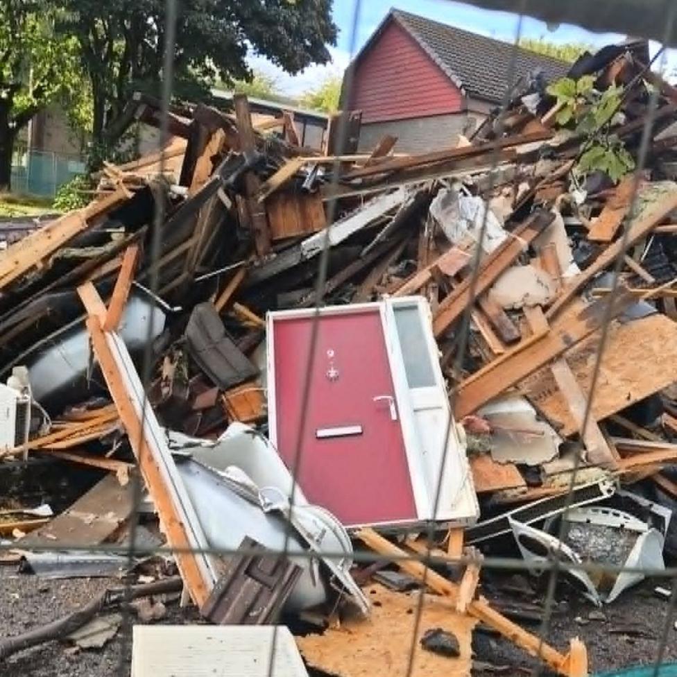 A demolished house behind a wire fence, the red front door is still intact, lying among the remains of the building.