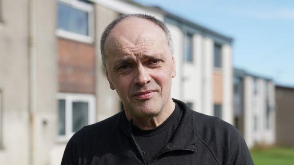 David Porter, a man looking at the camera, he is wearing a black top and is standing in front of housing.
