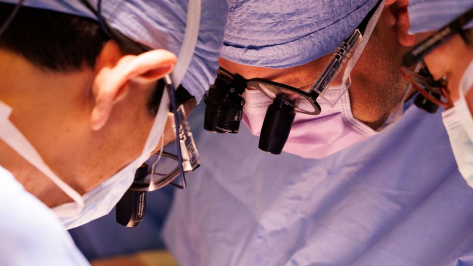 A tightly framed photograph shows several members of a surgical team gathered closely during a procedure. Each person is wearing blue surgical clothing, face masks, and magnifying lenses mounted on protective eyewear. The image focuses on their faces and equipment as they lean inward and look down toward an area outside the frame. Bright operating-room lighting illuminates the scene, while the background remains softly blurred in shades of blue. The composition emphasises concentration, precision, and the collaborative nature of a medical procedure.