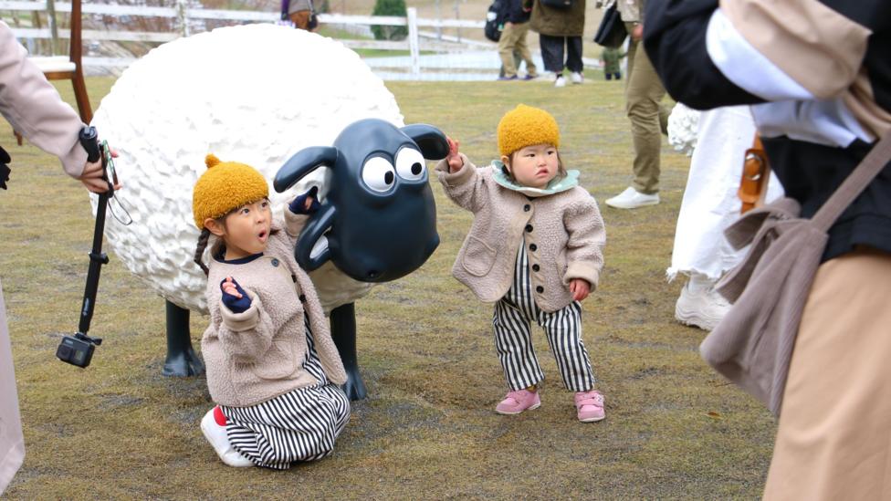 Two small Asian children are posing with a Shaun the Sheep statue. They are wearing matching outfits with yellow hats, beige fleece jackets and matching stripey trousers.