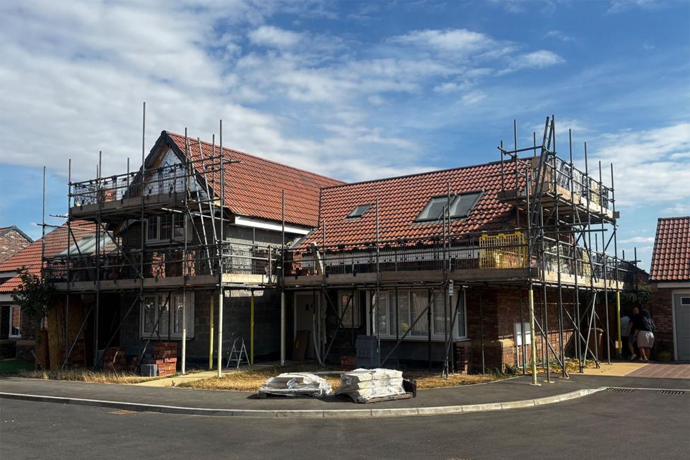 A two‑storey house stands surrounded by scaffolding, with red roof tiles and skylights visible. Construction materials, including wrapped supplies and stacks of bricks, sit in front of the partially finished exterior under a cloudy daytime sky.