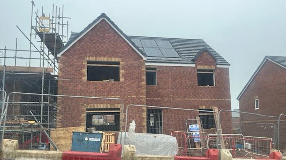 A two‑storey brick house under construction with dark roof tiles and solar panels. Open window and door spaces show the interior unfinished. Scaffolding stands on the left side, while pallets, barriers and wrapped materials sit behind metal fencing at the front.