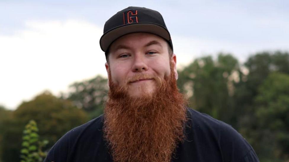 Luke has a long reddish‑brown beard and stands outdoors wearing a black baseball cap with an orange “G” and a dark shirt, set against green trees and a cloudy sky.